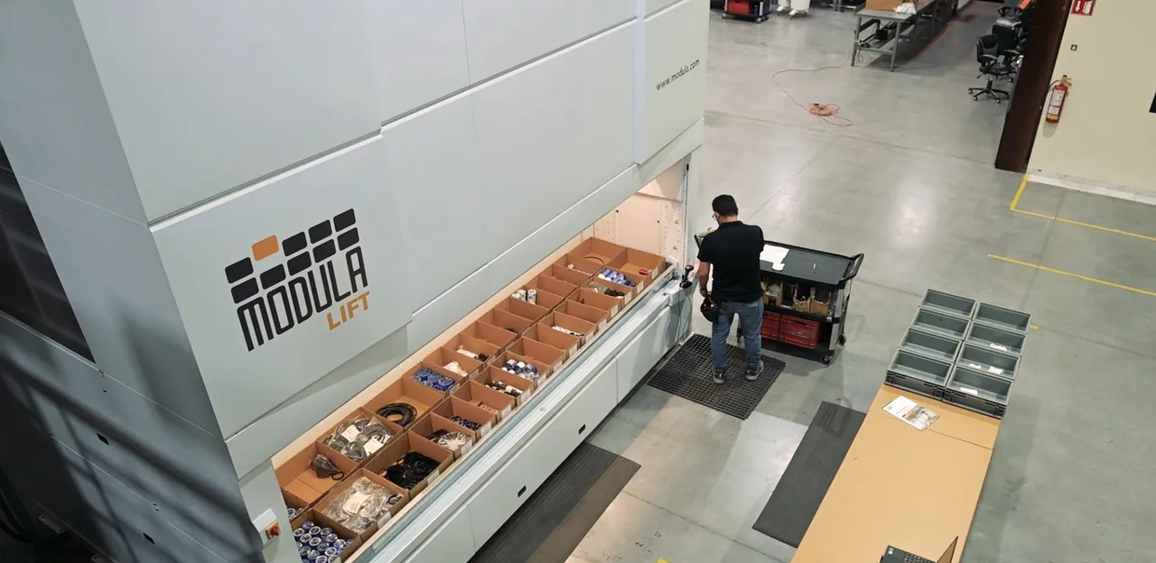 Vista desde arriba de la bahía de un Modula Lift en La Capital: un operador con terminal toma piezas de una bandeja con cajas de sellos; fotografía de la ficha oficial del caso