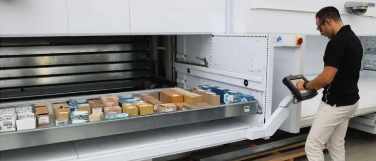 Operador con una terminal en la mano frente a la bahía de un almacén vertical Modula; la bandeja extendida lleva cajas de pedidos ordenadas por compartimento